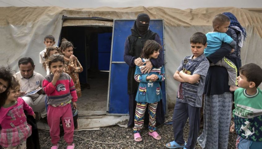 A group of children around a woman in a burqa; standing outside a refugee camp