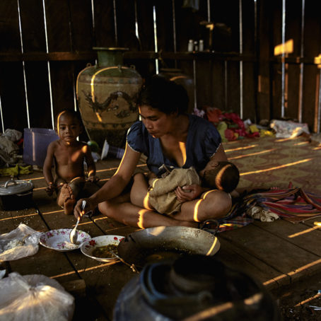A mother has a baby on her lap and another young child besides her in a hut in Cambodia, she is feeding her sons some food.