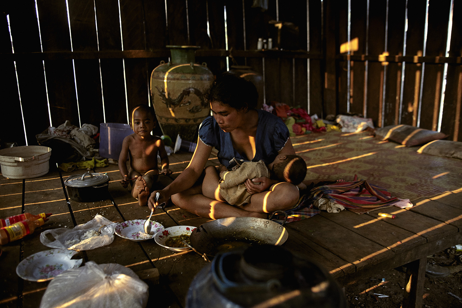 A mother has a baby on her lap and another young child besides her in a hut in Cambodia, she is feeding her sons some food.