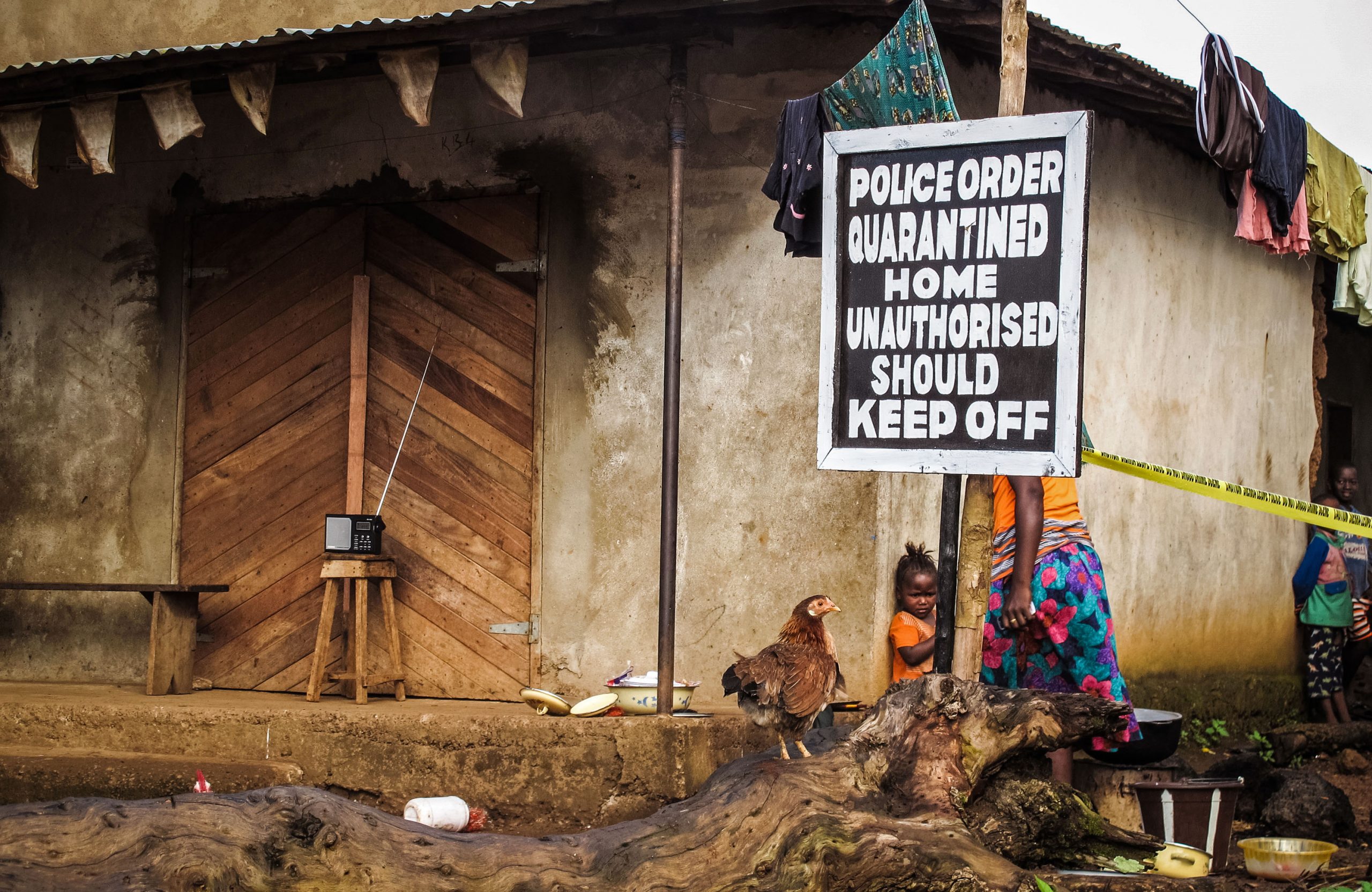 A young child with an adult and a rooster outside her home; a poster of police order quarantine warning outside the house.
