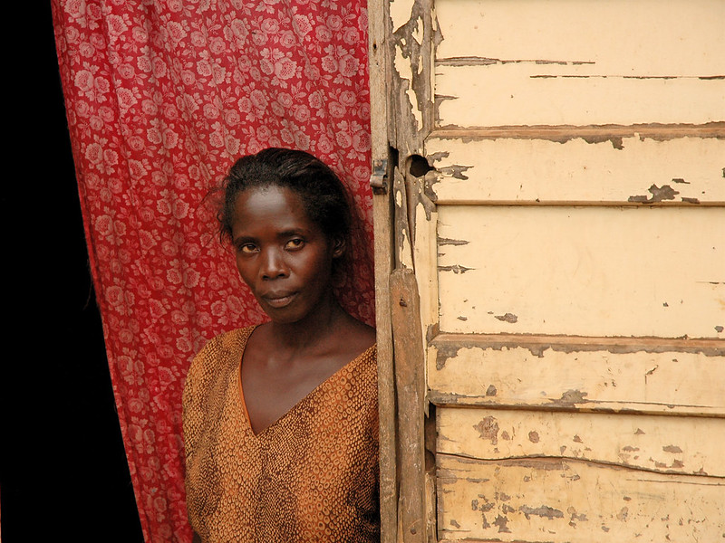 A Ugandan woman wearing a yellw dress, stands against a red backdrop at the door of her house