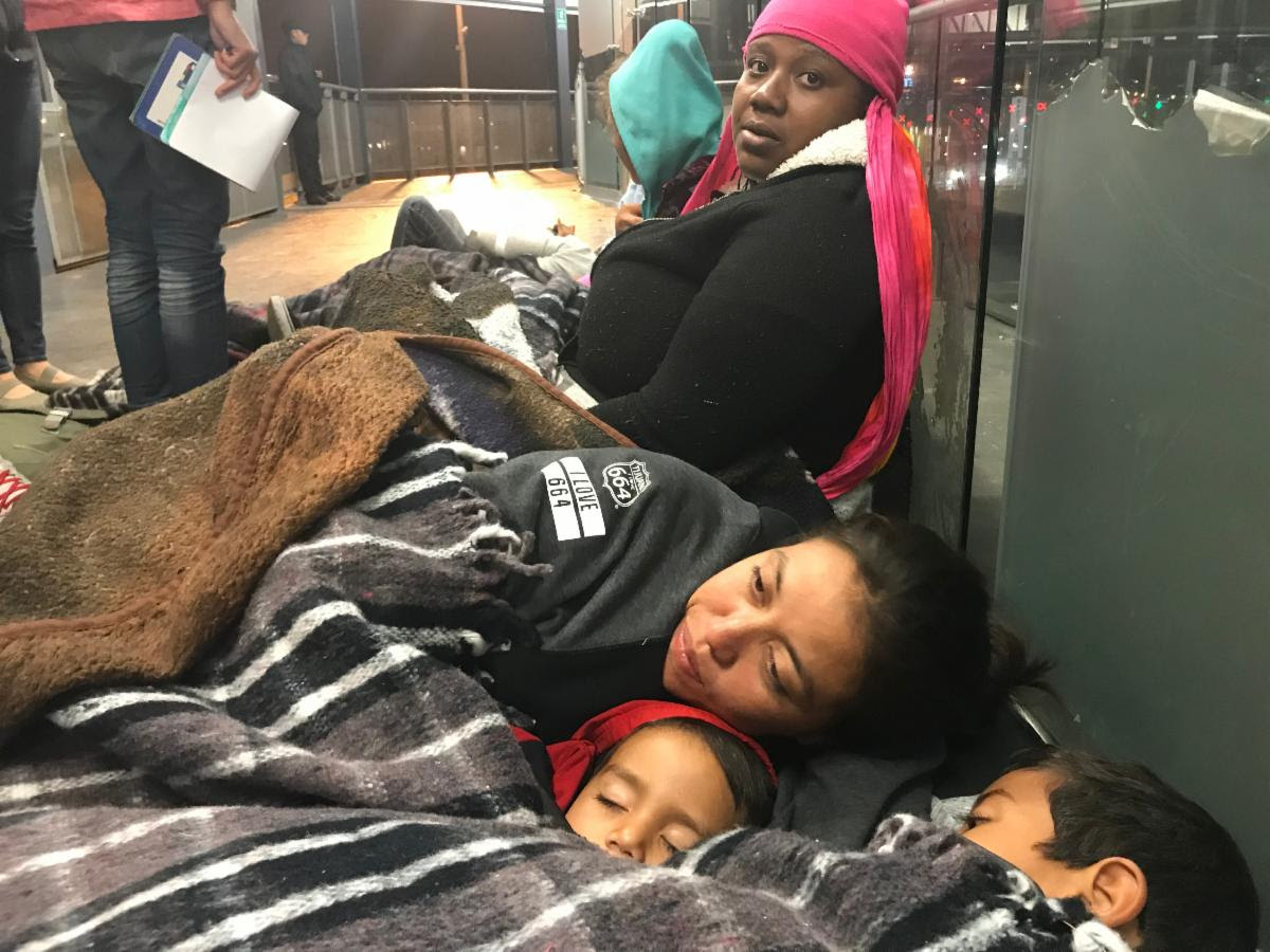 A woman wearing a pink pink sitting on the floor of a train station, another woman sleeps on the floor with two young children.