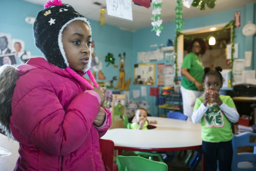Child wearing a pink winter jacket and a black hat in a childcare center; surrounded by more children and a childcare provider in the background.