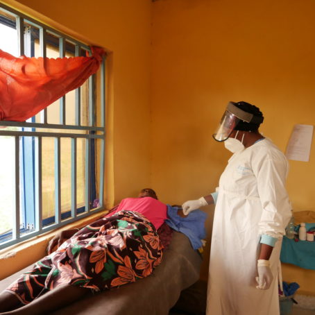 A nurse in full safety gear attends to a patient lying down on a bed nearby.