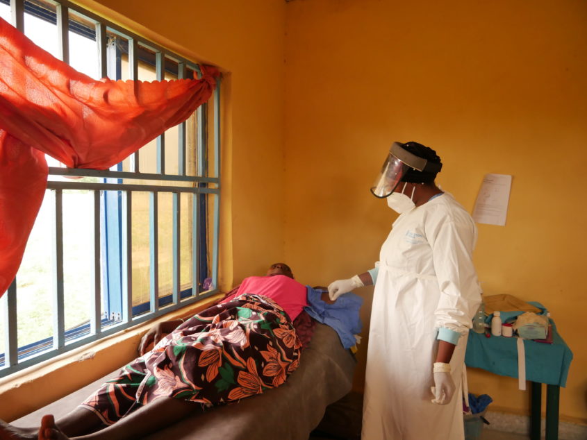 A nurse in full safety gear attends to a patient lying down on a bed nearby.