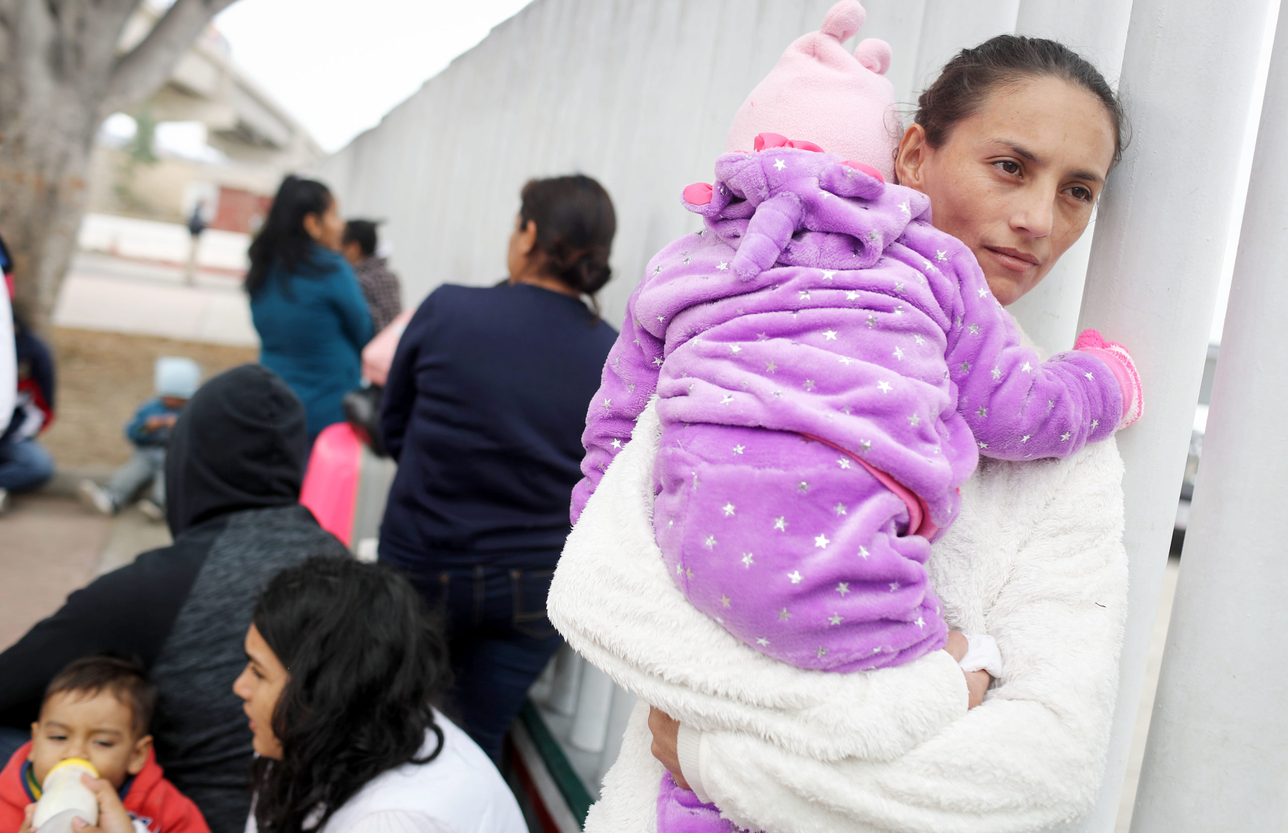 Woman holds her baby as she stands in line along a fence with other migrants