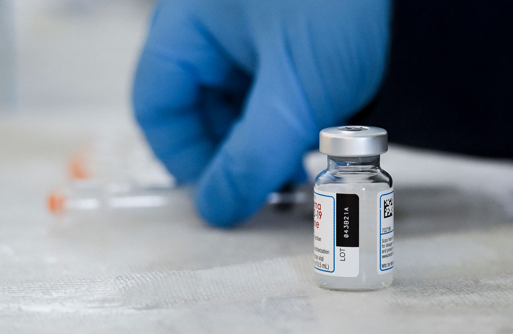 A picture of a vial with a vaccine in the foreground, with a person wearing a glove picking up a syringe in the background