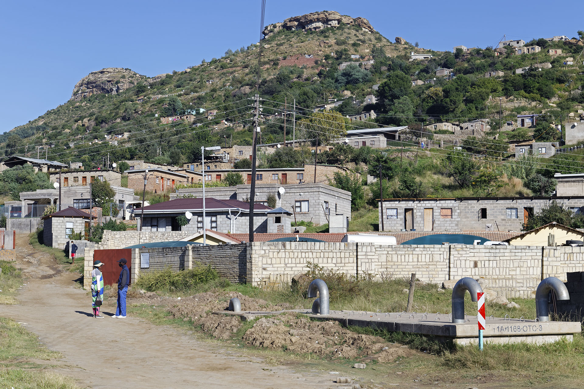 Photo of a village with a mountain and houses in the backdrop and a few people standing on the left side of the road