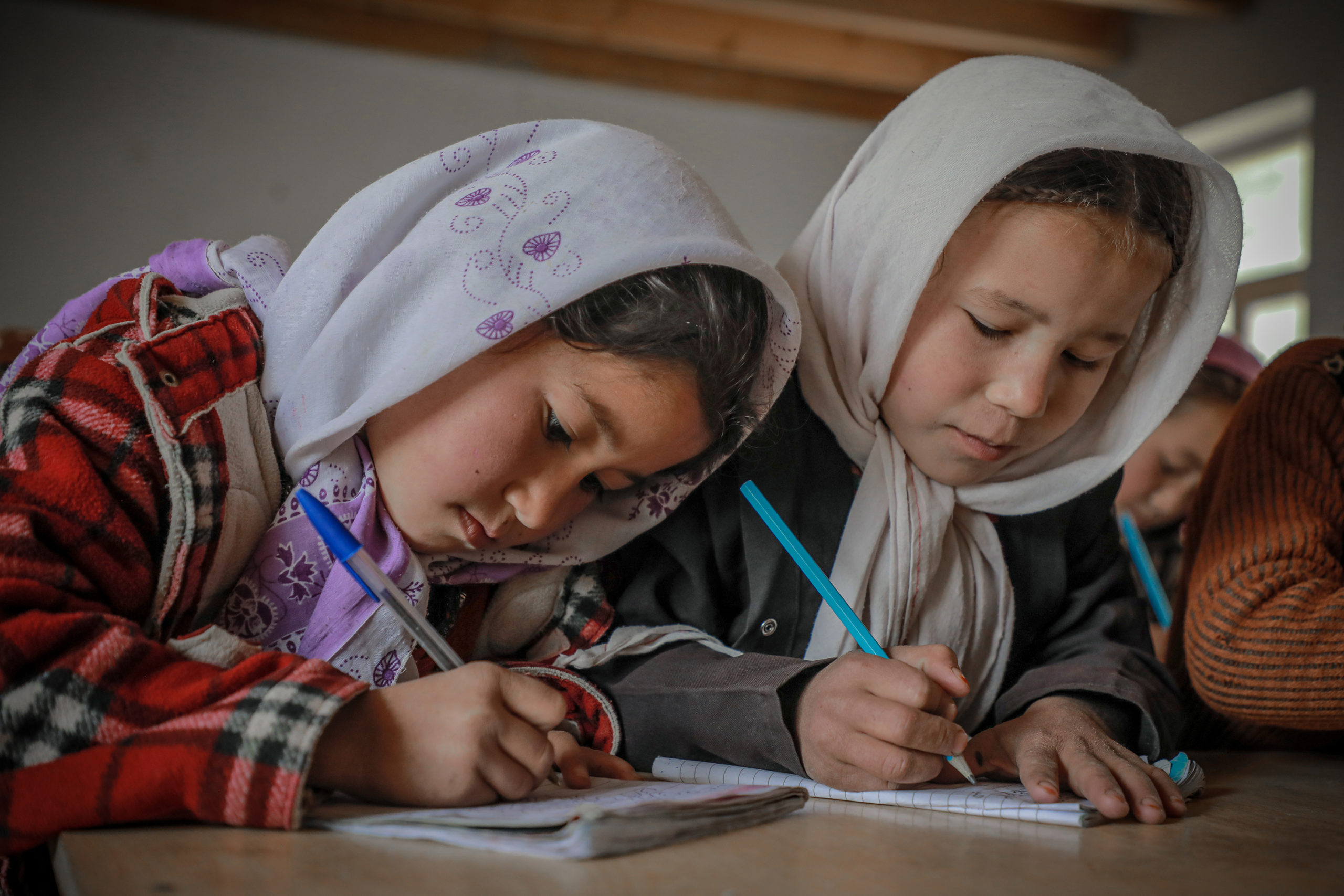 A photo of two school girls wearing headscarves writing in notebooks at a table
