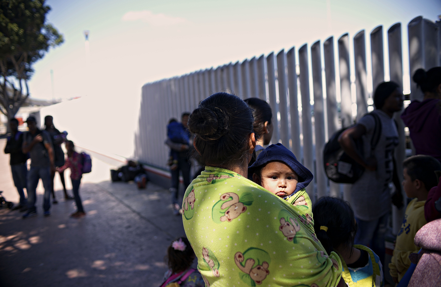 A photo of a woman's back as she holds a baby standing near a fence as others wait in line