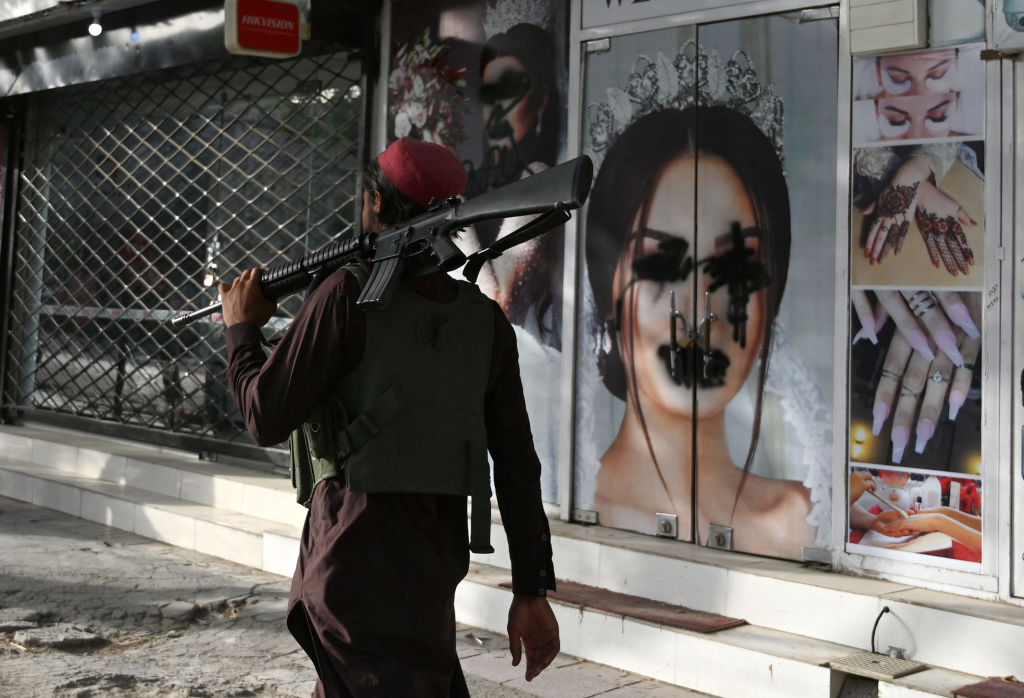 A photo of a man carrying a rifle walking past a defaced image of a woman
