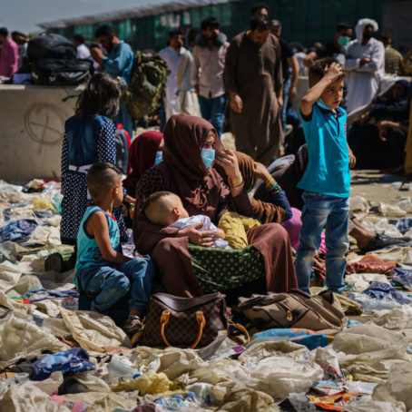 A photo of an Afghan woman holding a child as she sits with other children on a pile of trash and debris