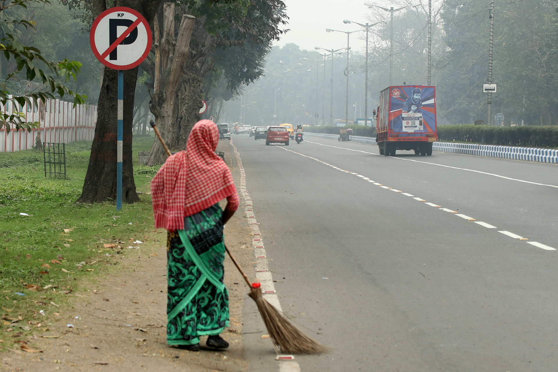 Photo of a woman sweeping a street while covering her face and head with a towel; smog in the background.