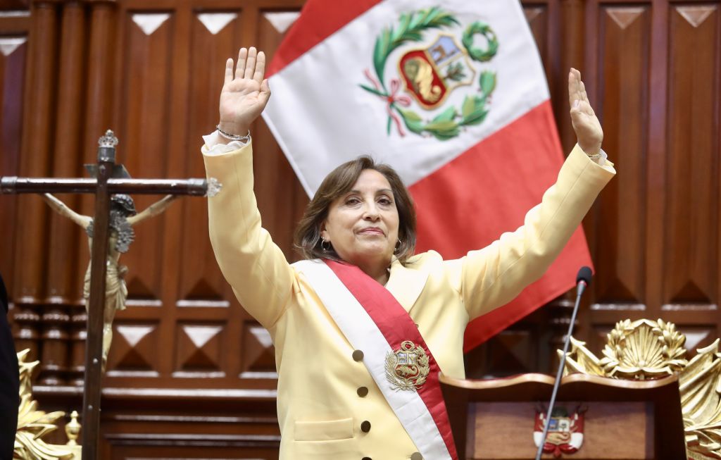 A woman wearing a sash with Peru's flag on it stands in a courtroom with a yellow suit and has her arms raised in the air