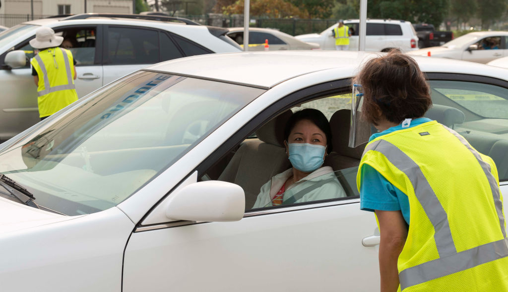 A woman wearing a face mask in a car talks to another woman standing outside of the car. 