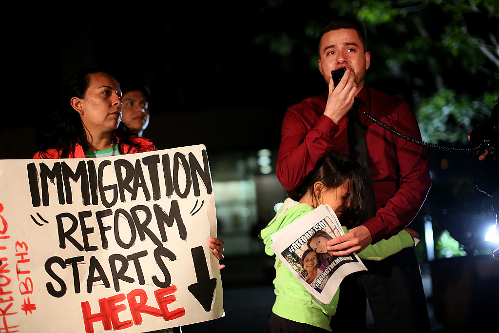 A man cries while holding a microphone and a girl hugs him. A woman is standing next to the guy with a sign that says "immigration reform starts here."