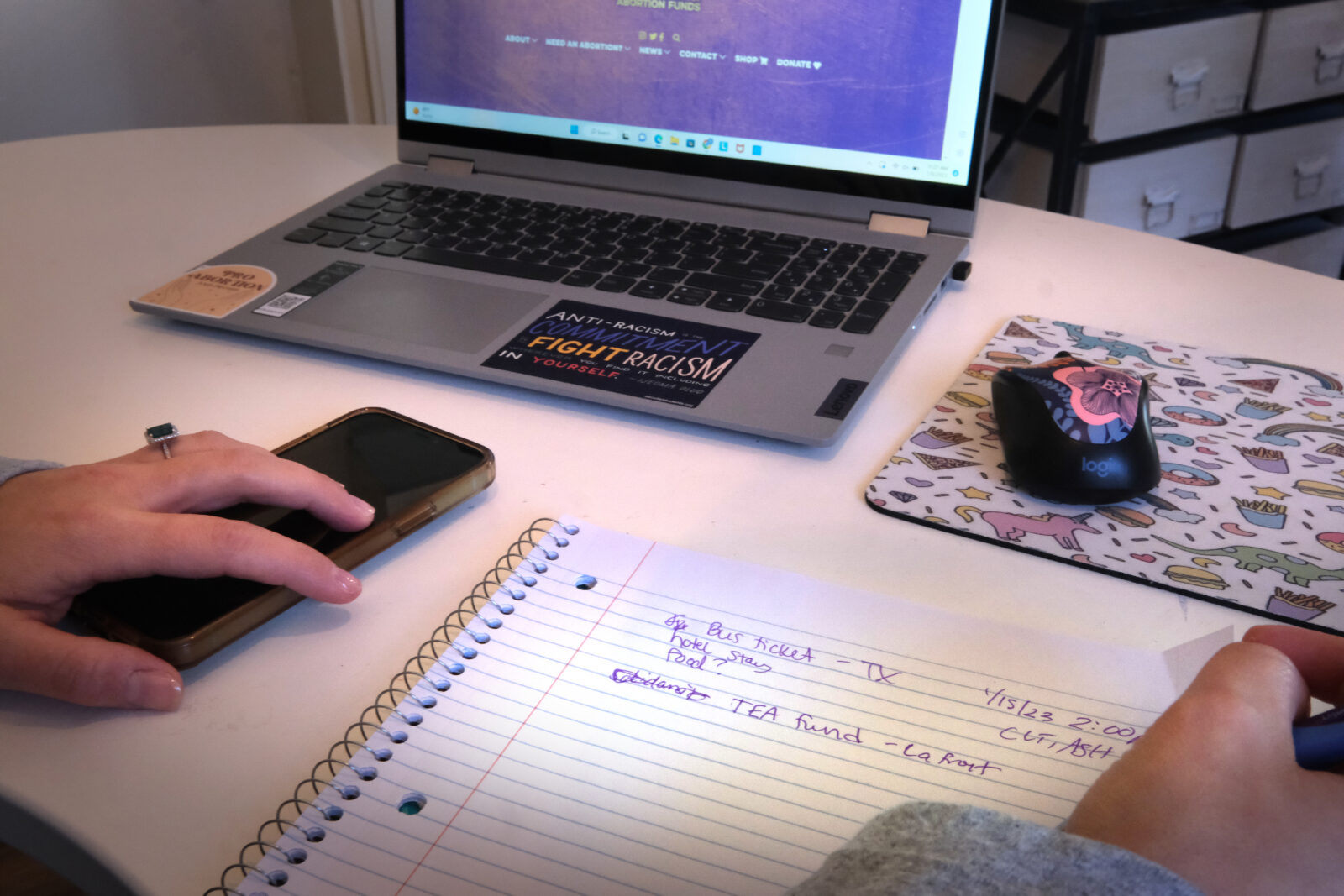 A note pad with purple writing on it, a laptop, computer mouse and cell phone sit on a white desk.