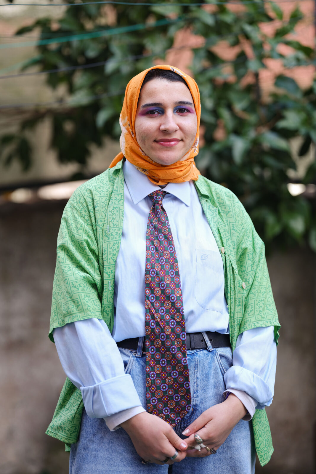 A woman stands wearing an orange head scarf with a green short sleeve shirt over a blue long sleeve shirt with a red and blue tie and jeans.