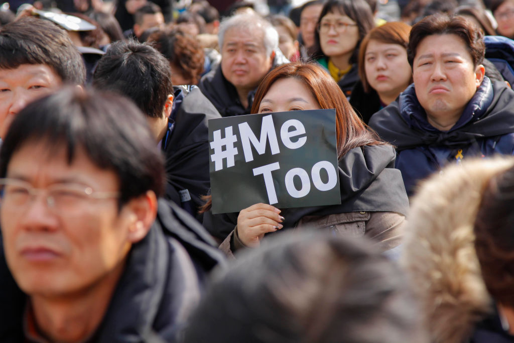 A woman covers her face with a #MeToo sign while sitting on the ground.