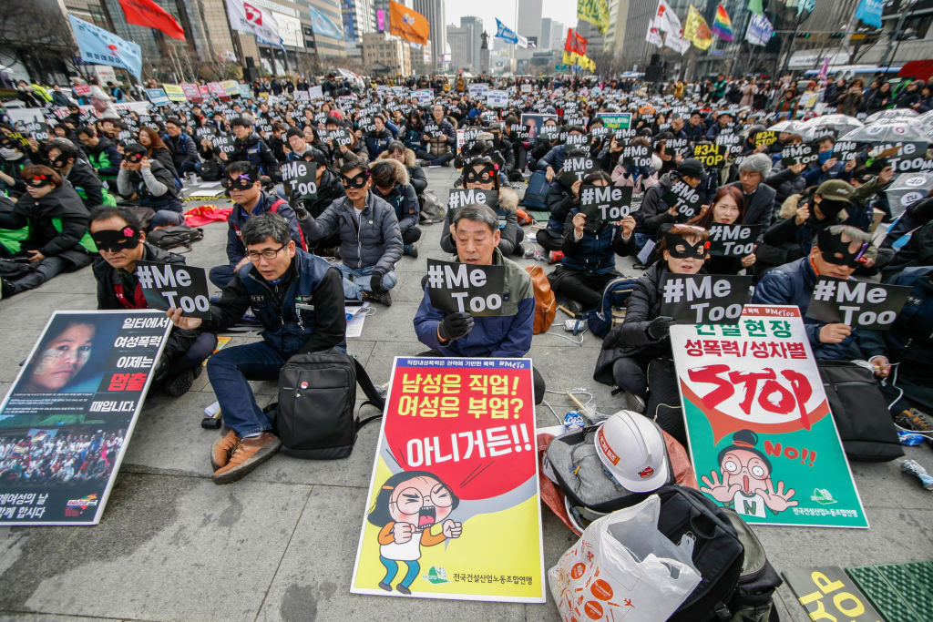 Hundreds of men and women in South Korea sit on the ground holding #MeToo posters.