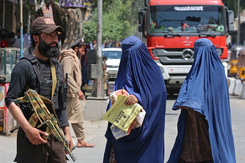 Two women wear blue burqas while a soldier stands next to them holding a rifle.