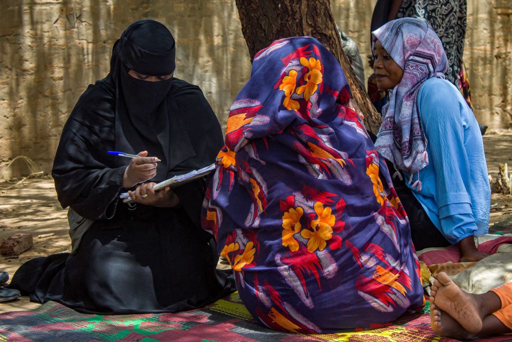 Three women sit on the ground. One woman writes on a notebook with a pen.